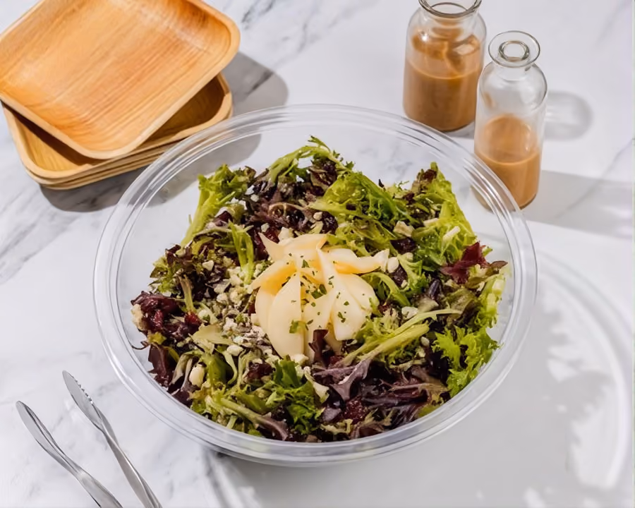 Mixed green salad with sliced pear in a clear delivery bowl, with dressing bottles and bamboo plates