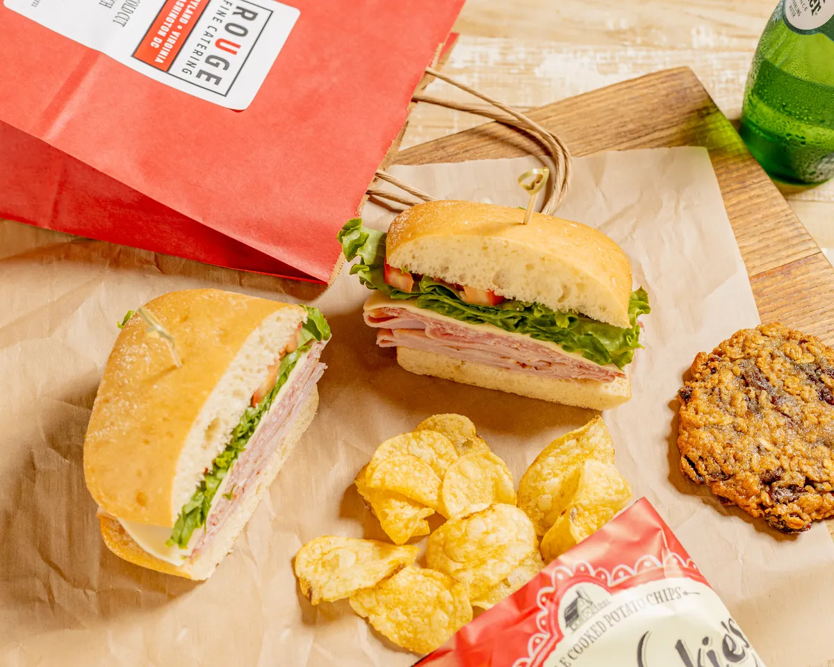 Boxed lunch with a ham and cheese sandwich, chips and a cookie beside a Rouge Catering bag