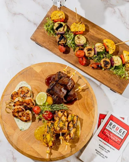Grilled shrimp, beef, chicken and vegetable skewers on wooden boards with a Rouge Catering card
