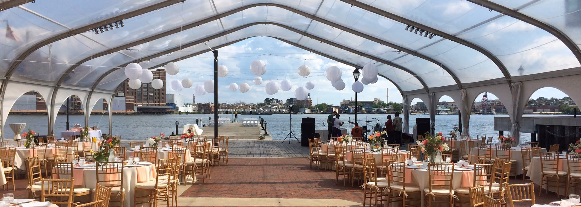 Waterfront tented reception with round tables, gold chairs and paper lanterns overlooking the harbor