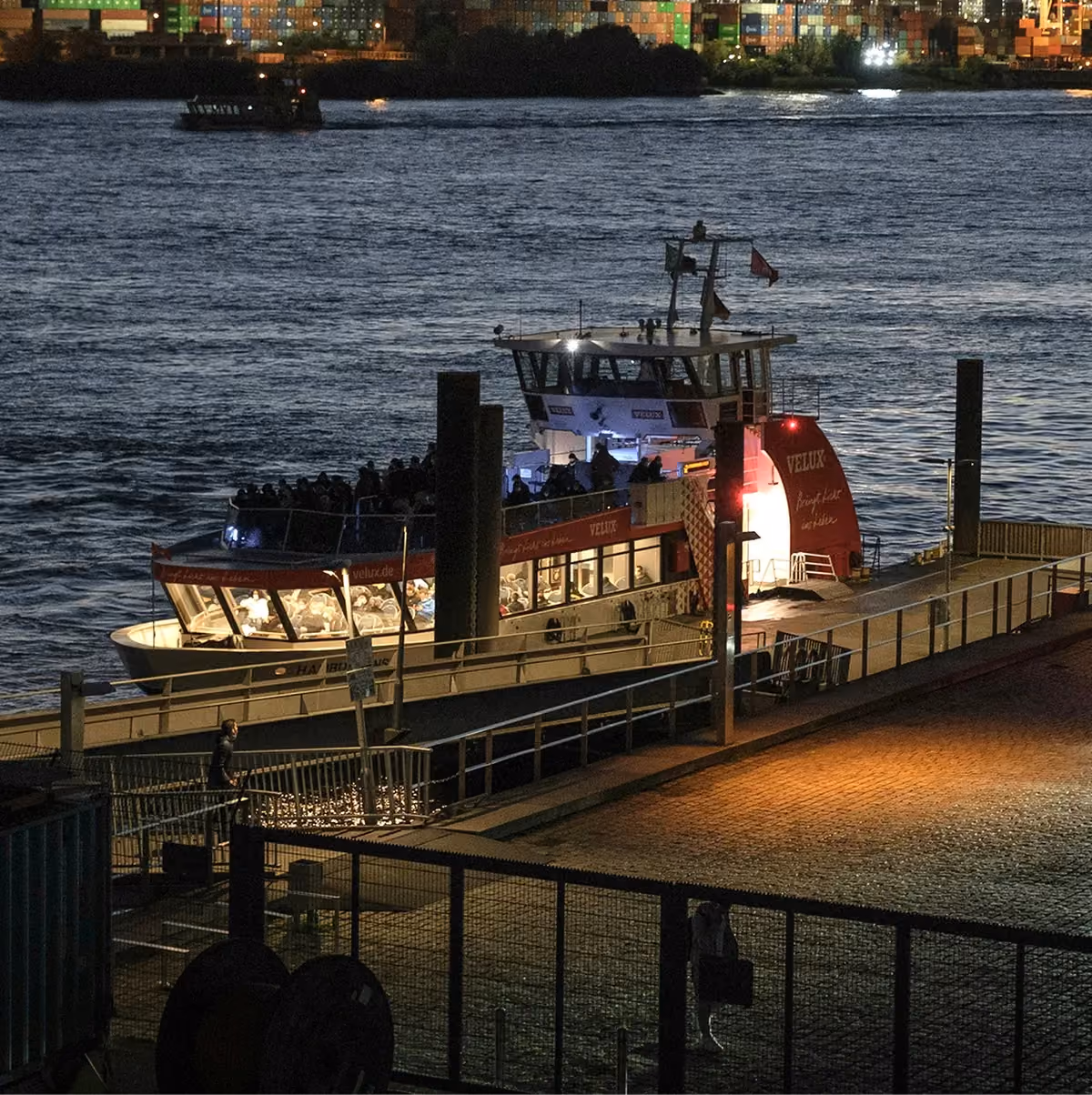 A passenger ferry moored at a harbour pier at dusk, with container cranes on the far shore