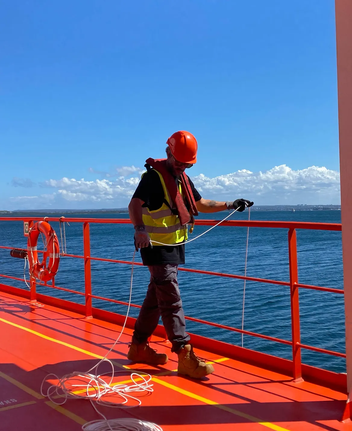 Crew member in a hard hat and hi-vis vest handling a tether line on a ship's red deck at sea