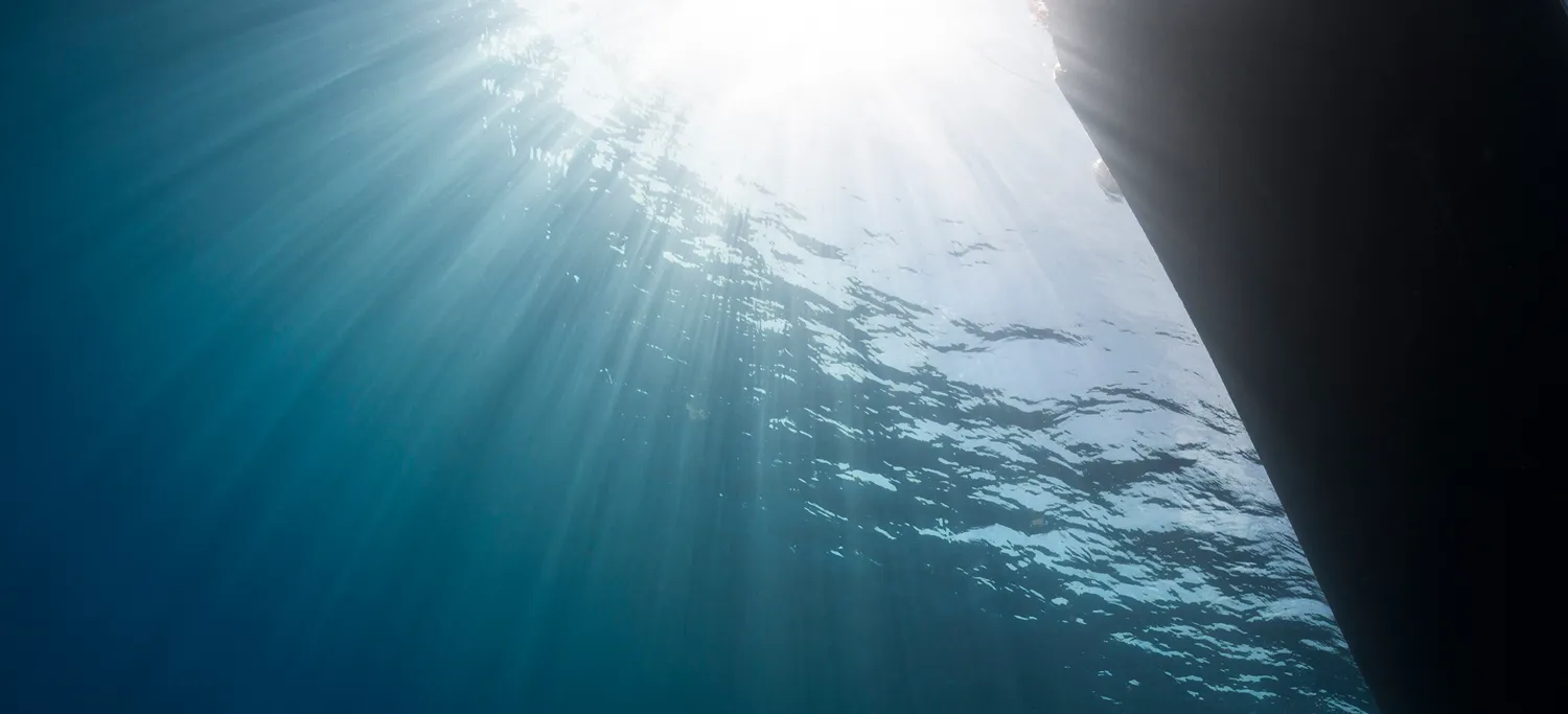 Sunlight shining through the water surface beside a ship's dark hull, seen from below