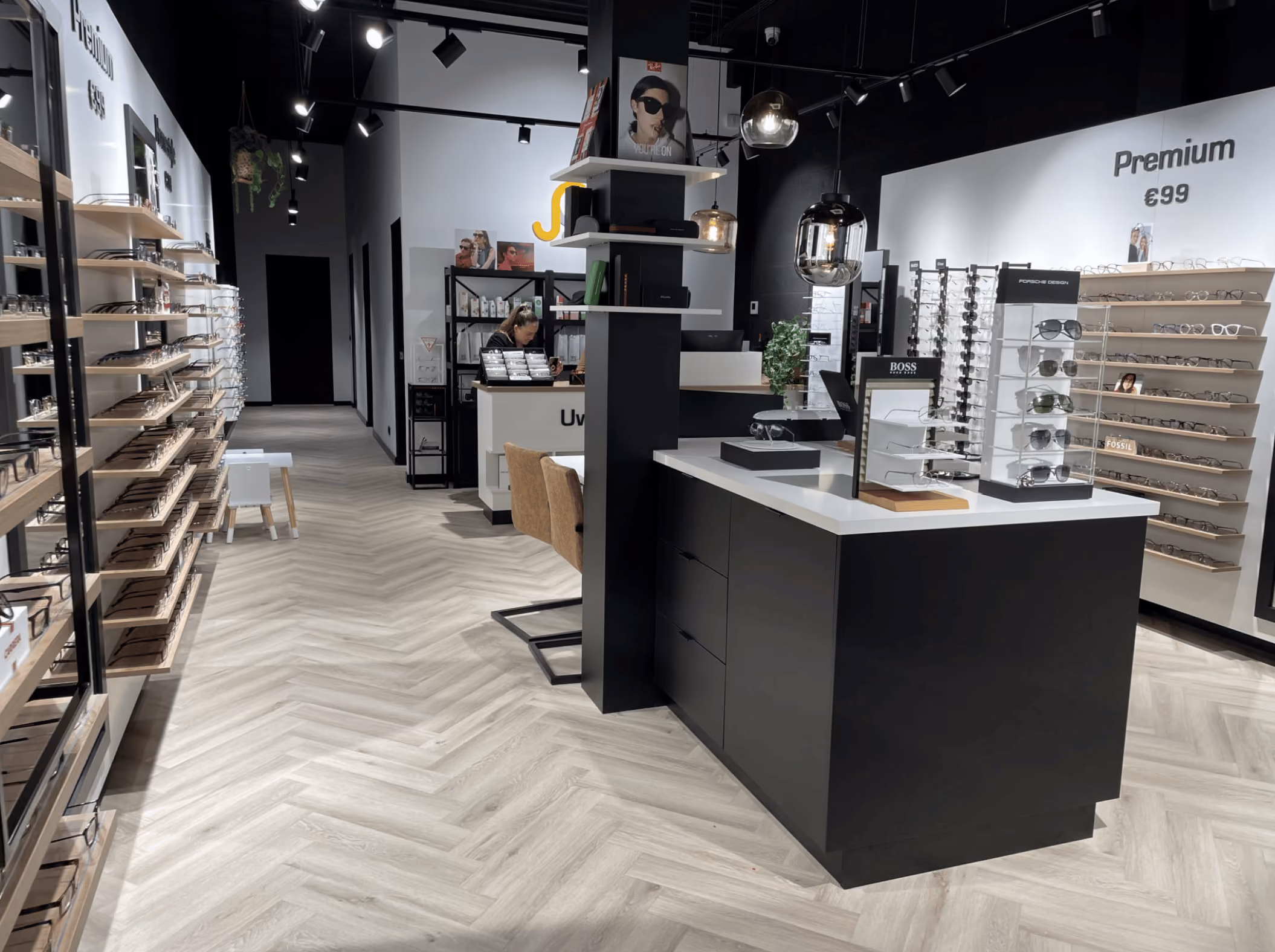 Moderne brillenwinkel met wandplanken en displays vol monturen, een centraal zwart en wit toonbankmeubel met merkbrillen, en een medewerker die achter de balie werkt.