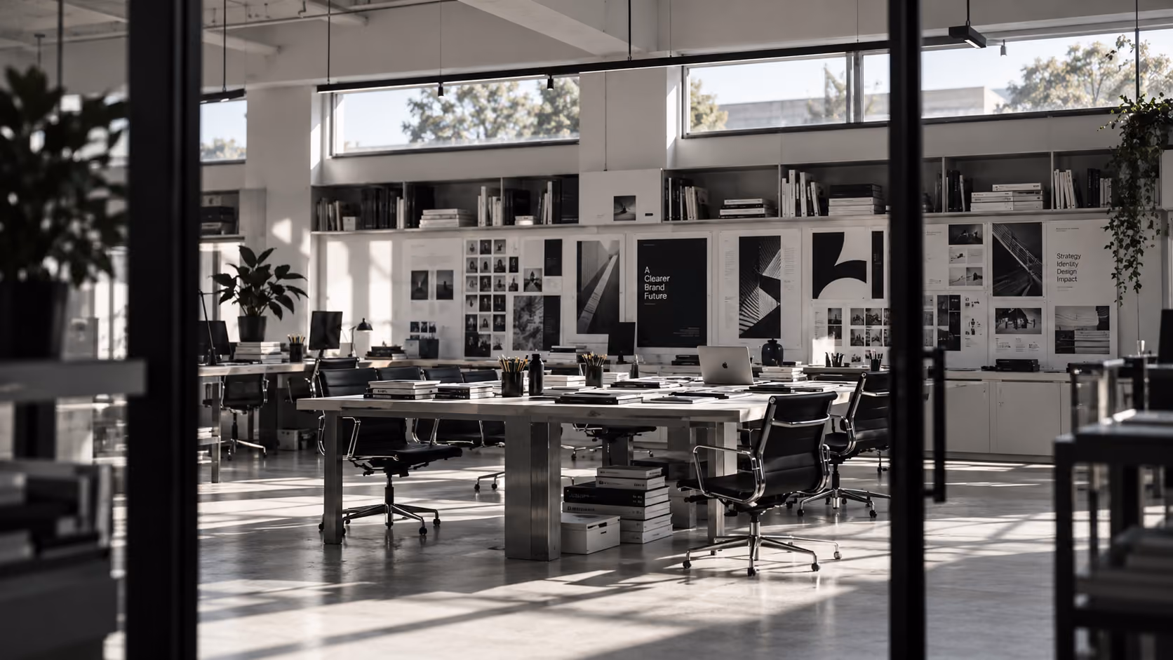 Moderne open kantoorruimte met grote ramen, een lange tafel met laptops, boeken en pennenbakken, zwarte bureaustoelen en planten, muren versierd met posters en foto's.