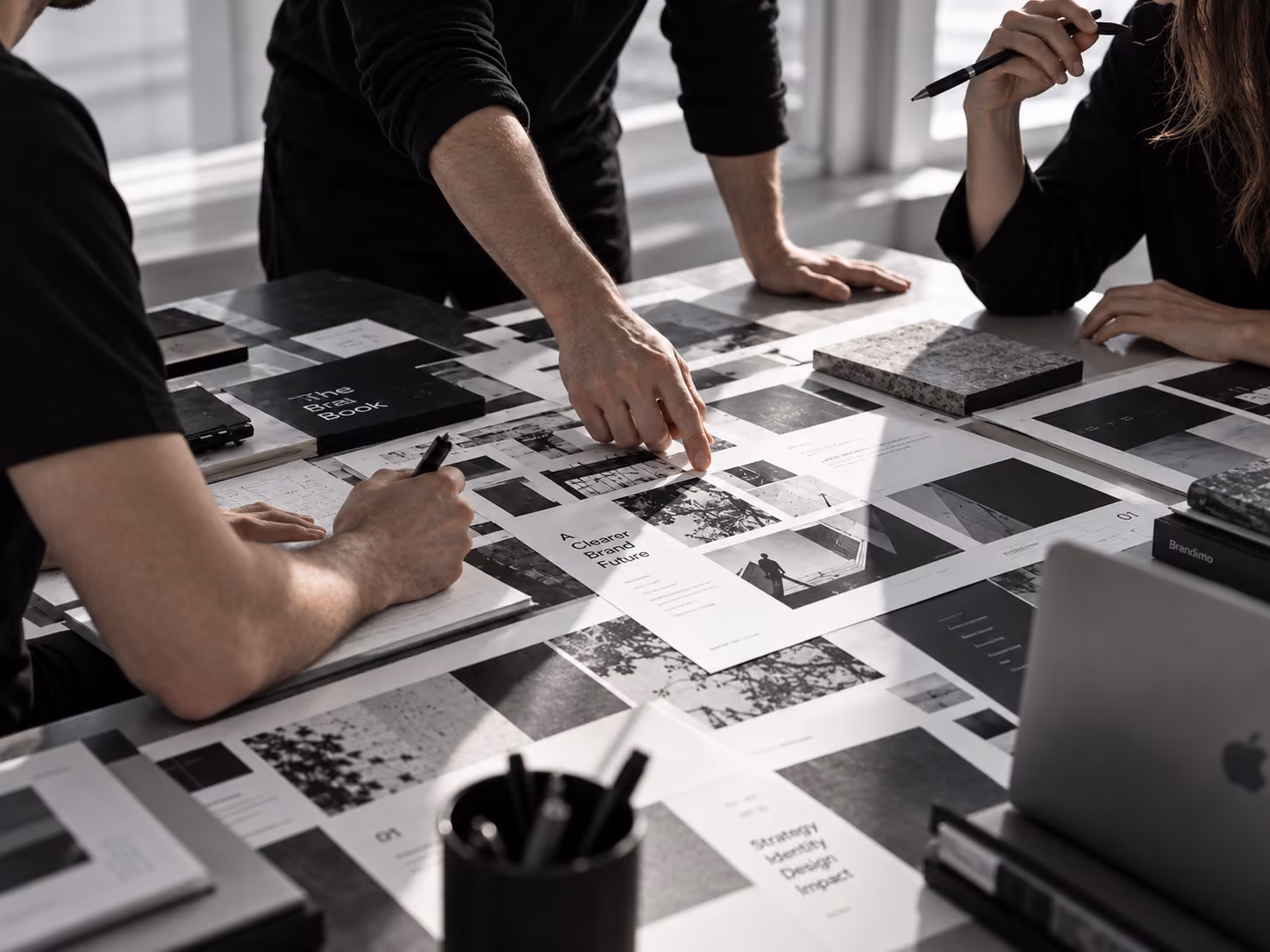 Drie mensen werken samen aan een tafel vol met geprinte pagina's en boeken over merkstrategie en design, waarbij een persoon wijst naar een document met de tekst 'A Clearer Brand Future'.
