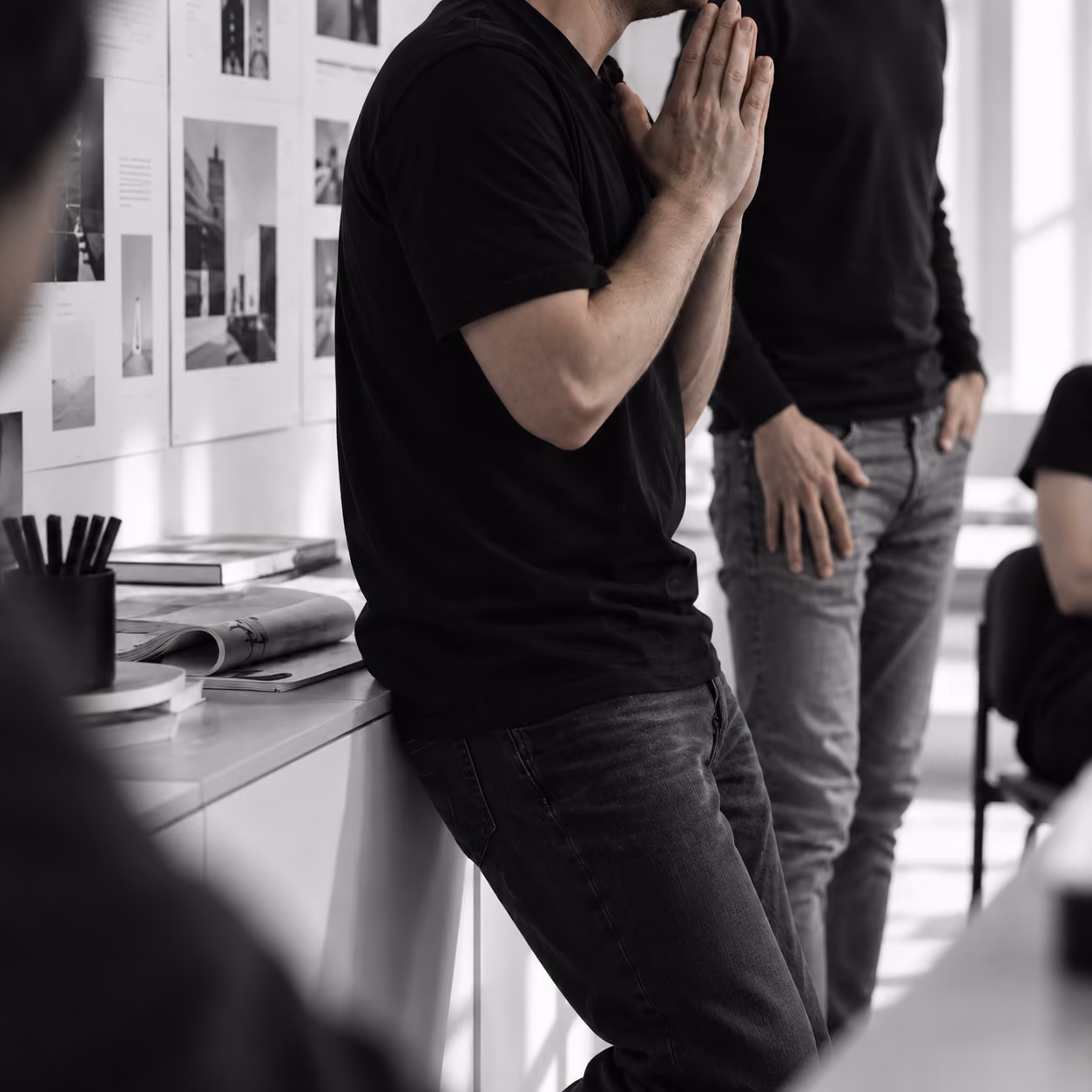 Twee mannen staan in een lichte, moderne kantoorruimte met zwart-witte foto's aan de muur; de ene man is gekleed in zwart en leunt tegen een kast terwijl hij zijn handen voor zijn gezicht houdt, de andere man staat naast hem met een hand in zijn broekzak.