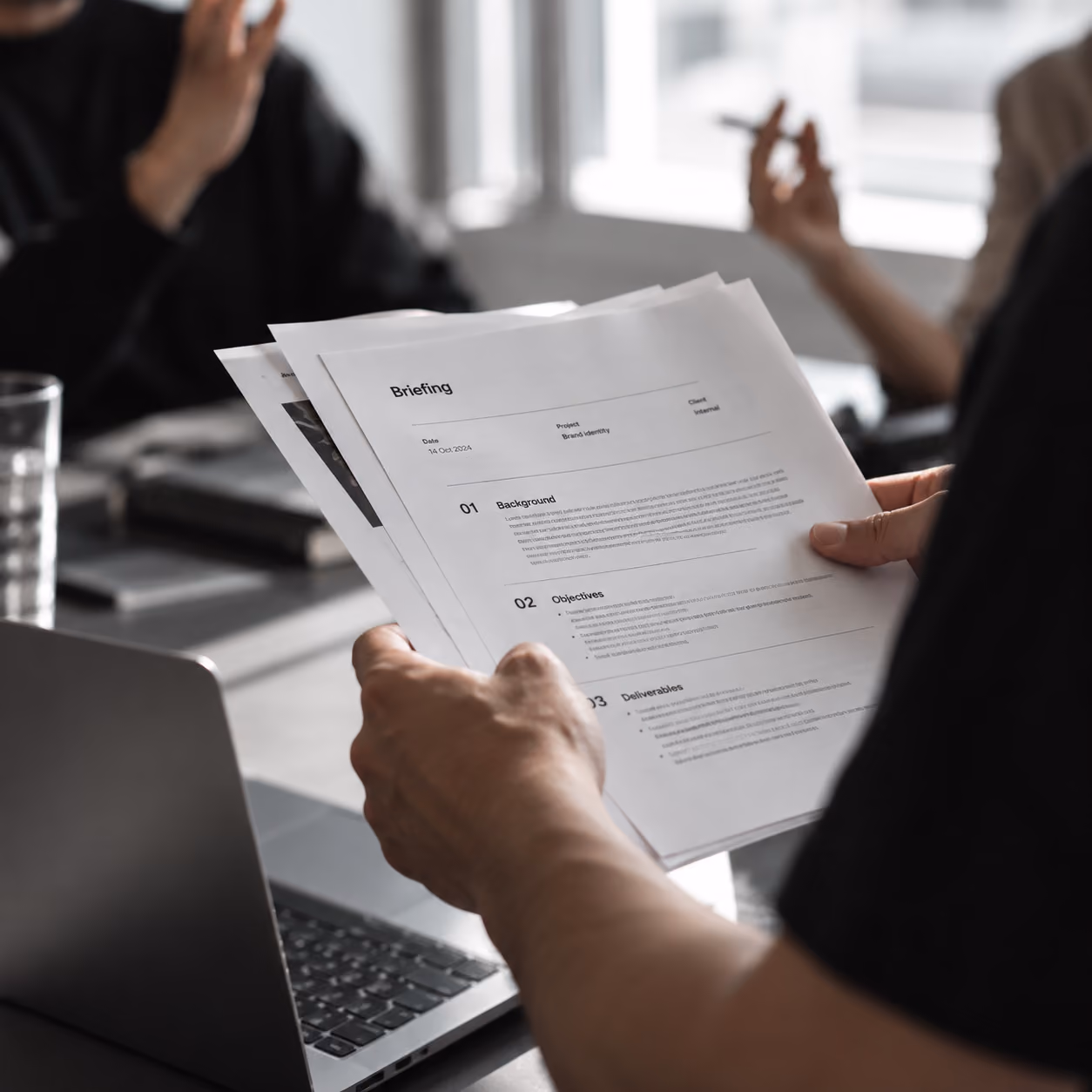 Iemand houdt documenten vast met de titel 'Briefing' in een kantooromgeving met op de achtergrond twee personen in gesprek, een laptop en een glas water.
