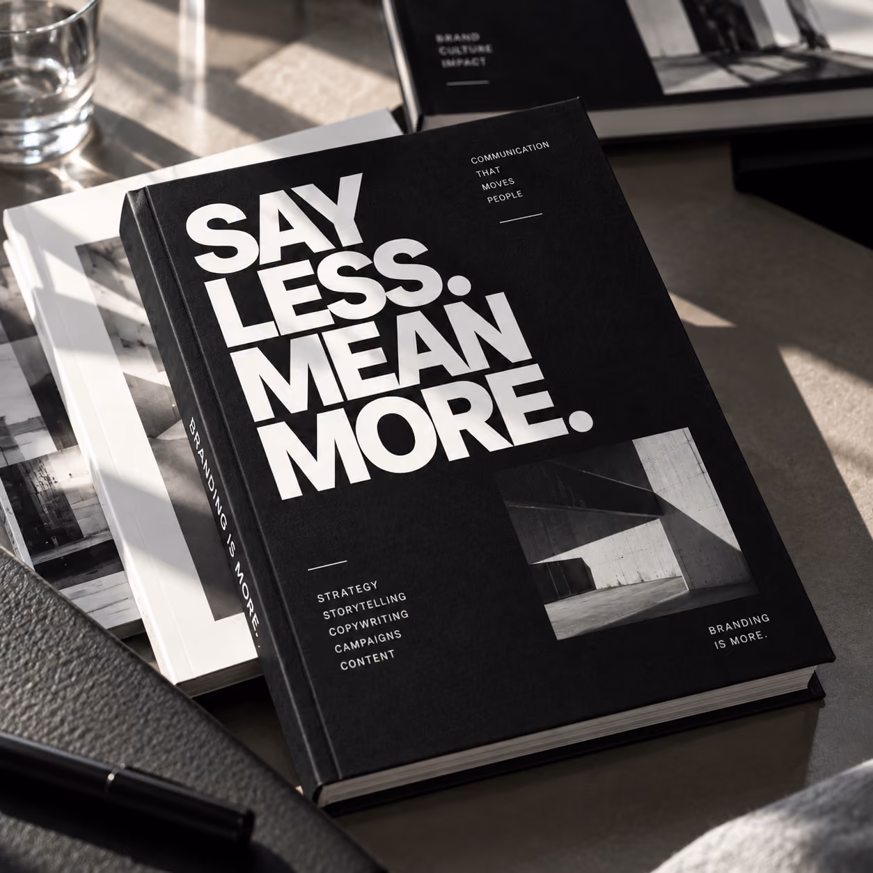 Een zwart boek met grote witte tekst SAY LESS. MEAN MORE. Andere tekst omvat branding-gerelateerde termen zoals strategie, storytelling, copywriting, campagnes en content; het boek ligt op een tafel naast een pen en andere boeken.