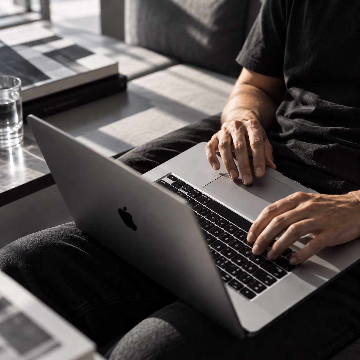 Persoon die op een MacBook-computer werkt, zittend op een bank met zonlicht dat schaduwen werpt op de achtergrond, met boeken en een glas water op een tafel naast hen.