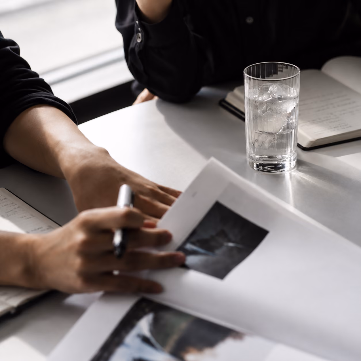 Twee mensen zitten aan een tafel met notitieblokken; een persoon houdt een pen en bladert door een document met foto's, terwijl een glas water met ijsblokjes op de tafel staat.