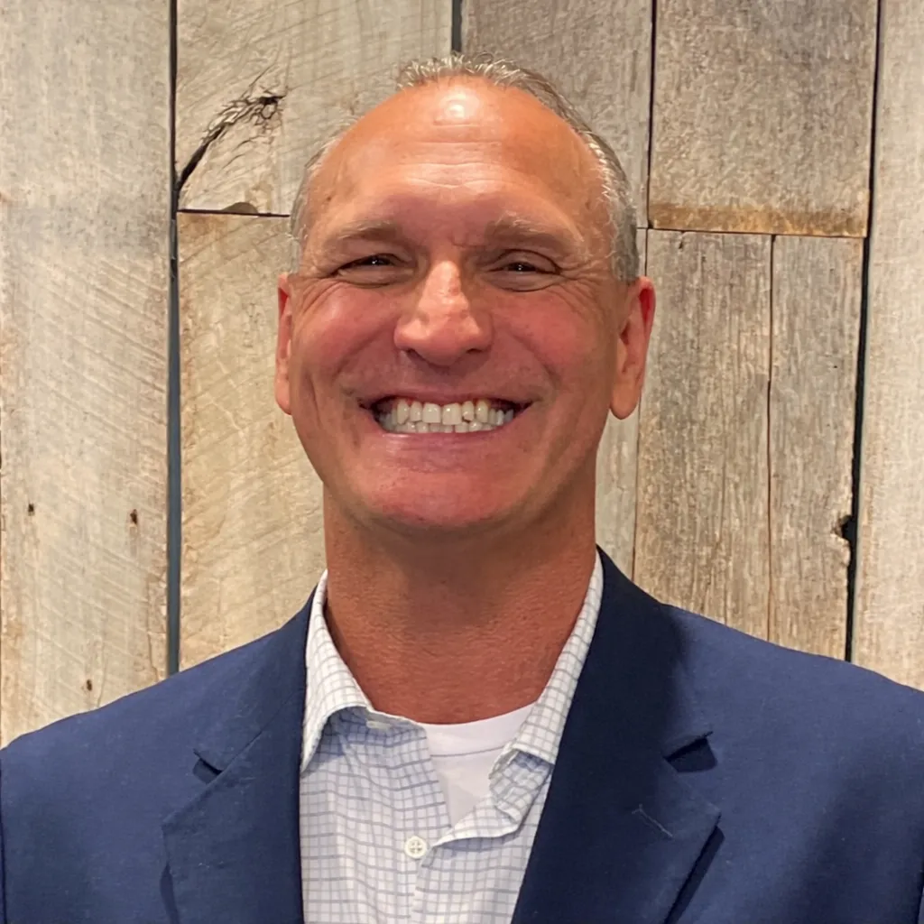 Smiling middle-aged man in a blue blazer and checked shirt standing in front of a wooden plank wall.