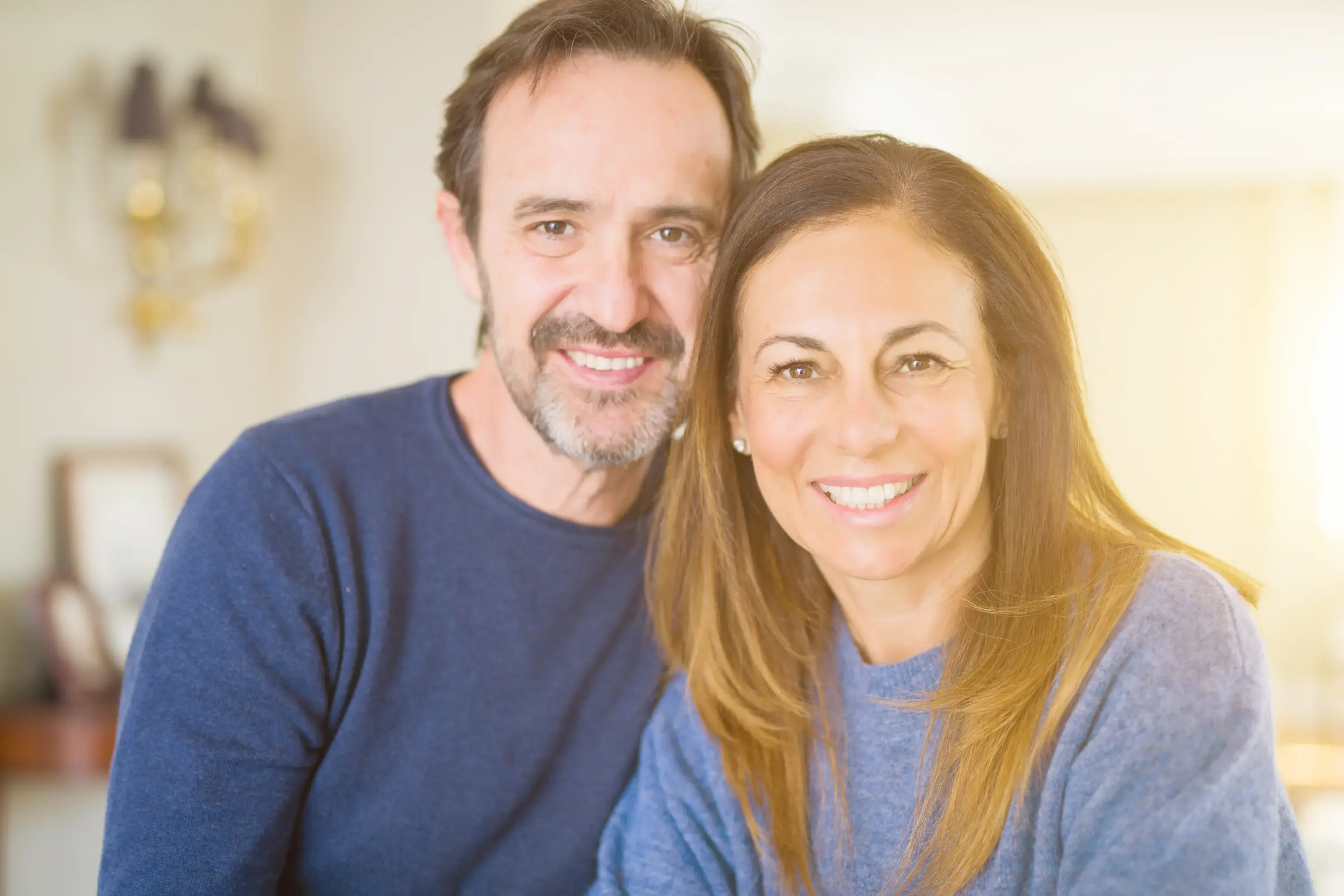 Smiling middle-aged couple wearing blue sweaters posing closely indoors with soft sunlight in the background.