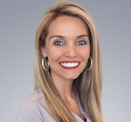 Smiling woman with long blonde hair, wearing silver hoop earrings and a light gray top, against a plain gray background.