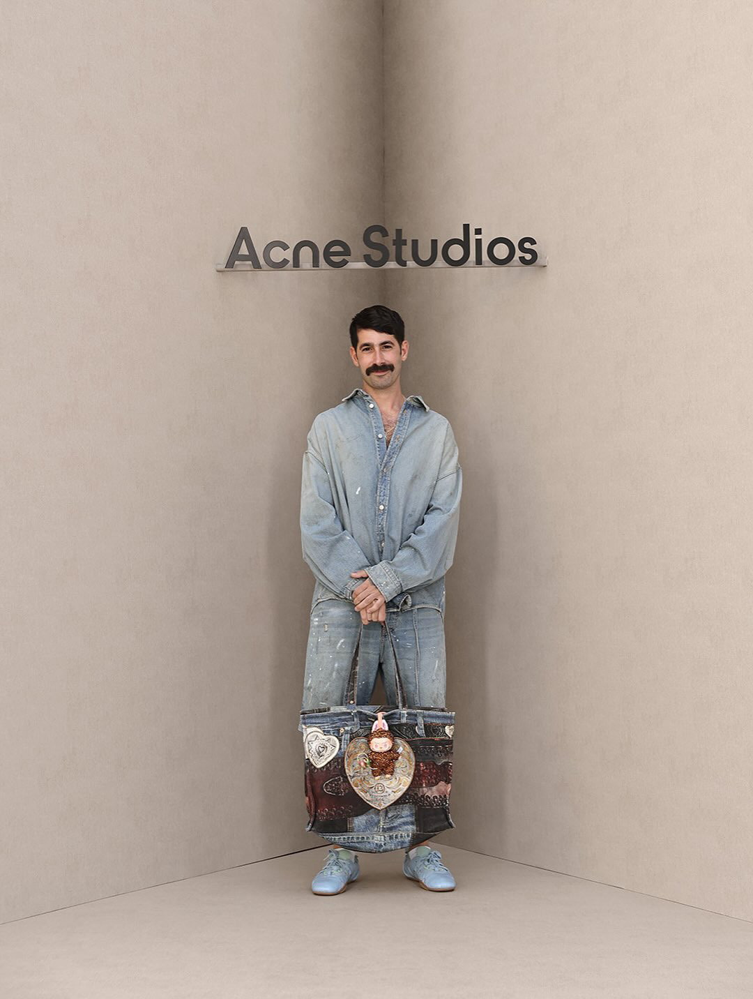 Man in denim holding a patterned tote bag in front of an Acne Studios sign, SS26 photocall.