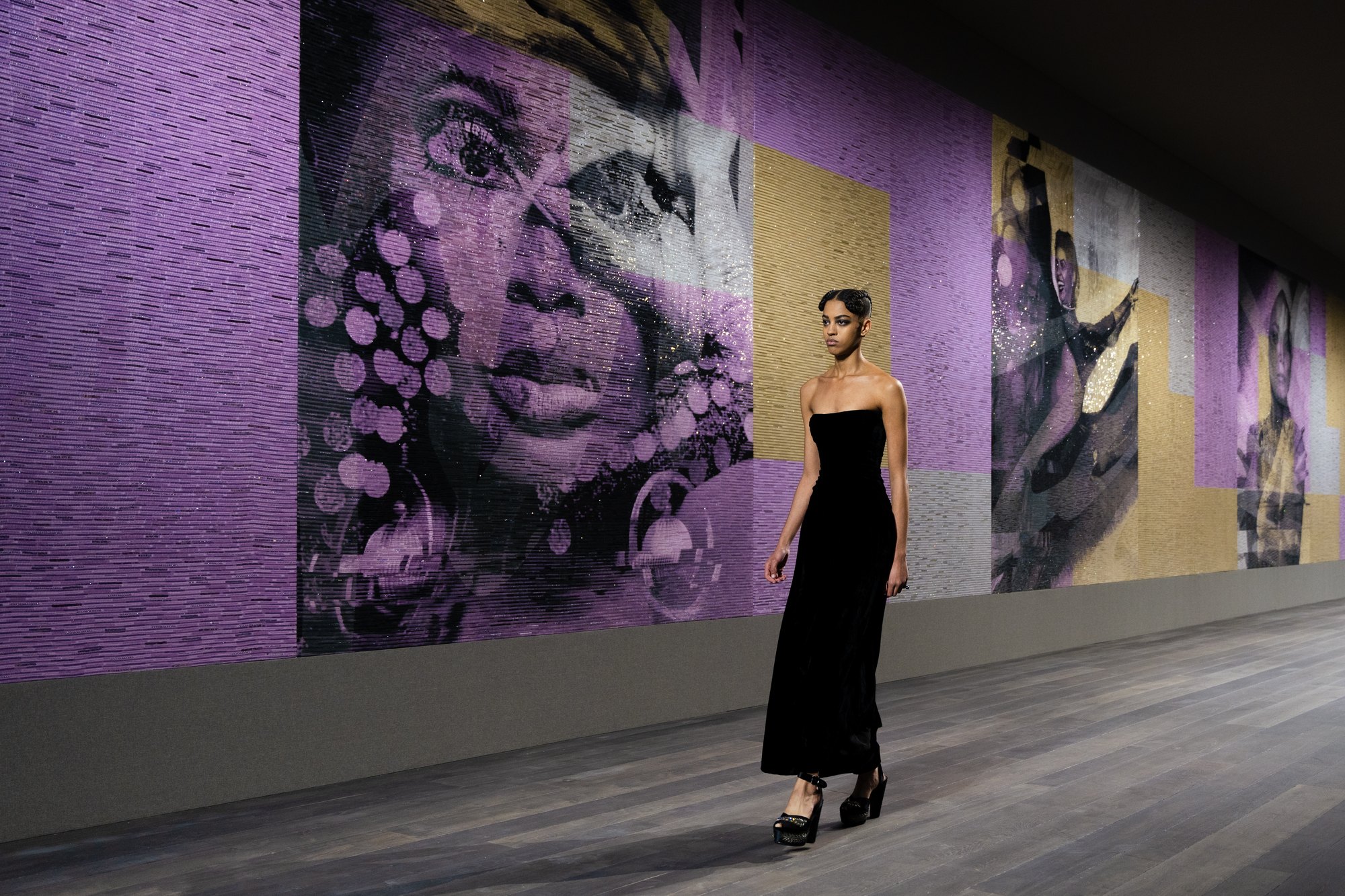 Model in a black dress walking in front of a wall printed with large portrait artworks, Dior Couture SS23, Musée Rodin.