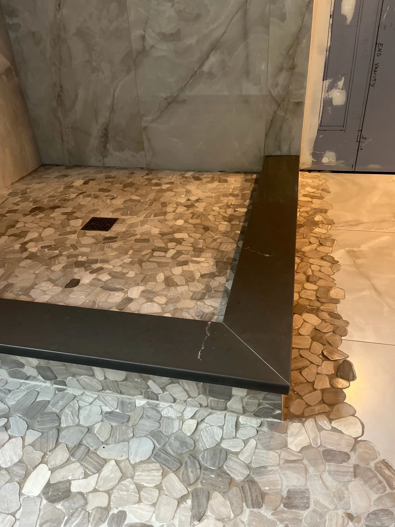 Custom-cut mosaic shower transition — Pebble mosaic meets adjoining tile along a shaped edge. A dark curb cap finishes the shower entry.