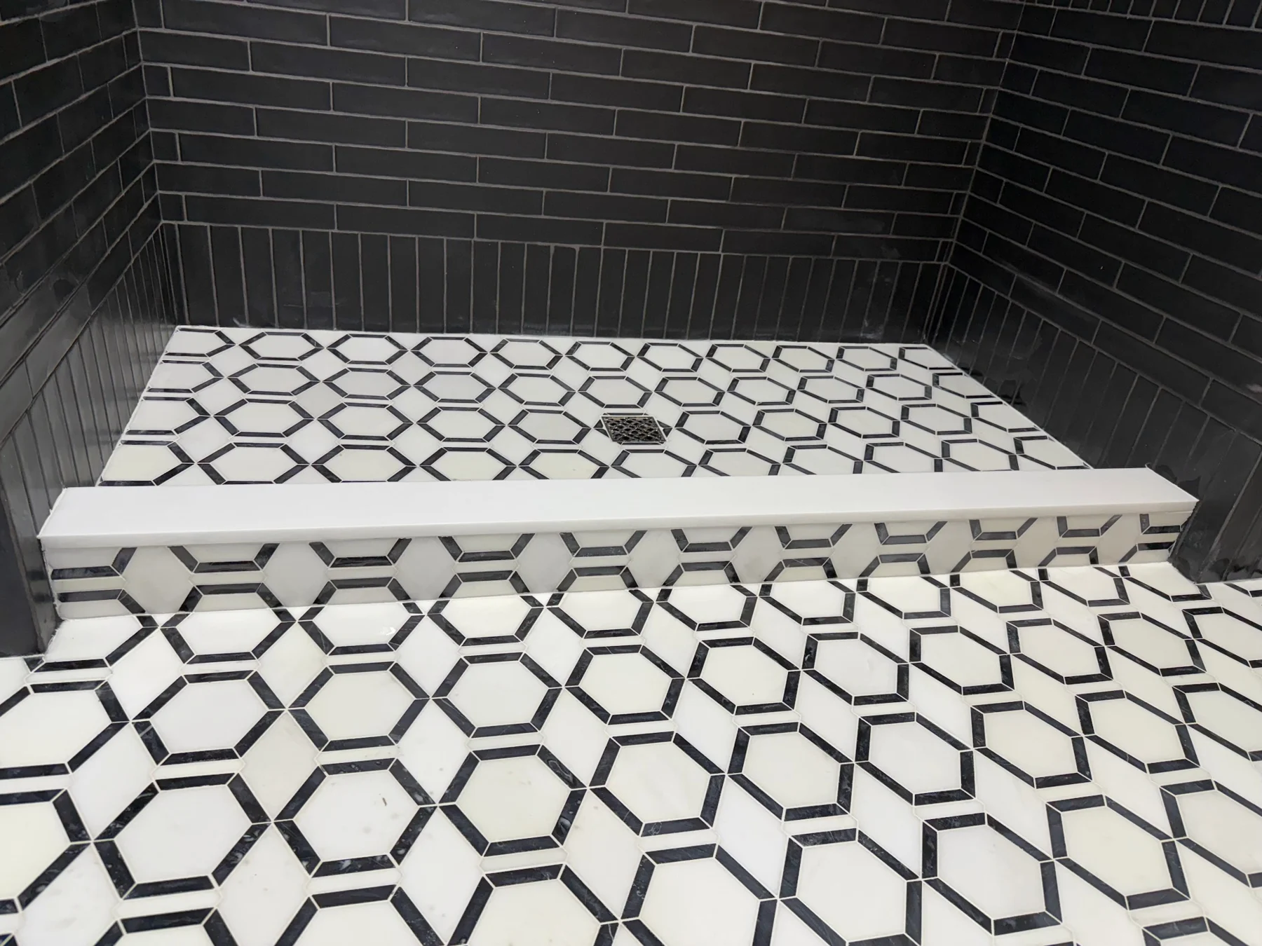 Geometric mosaic shower floor — Black-and-white mosaic continues across the shower floor and curb face, with a contrasting curb cap.