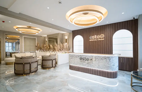 Modern medical group reception area with a marble front desk displaying 'este Medical Group' logo, curved chairs, decorative pampas grass, warm circular ceiling lights, and wood-paneled walls with arched niches.
