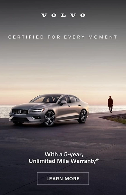 Silver Volvo sedan parked on a paved area near water during sunset with a person walking in the distance; text above reads 'VOLVO CERTIFIED FOR EVERY MOMENT' and below 'With a 5-year, Unlimited Mile Warranty*' with a 'LEARN MORE' button.