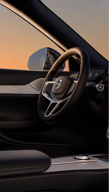 Interior view of a Volvo car showing a close-up of the steering wheel with the Volvo logo, part of the dashboard with a digital display, a side mirror, and a center console with wood trim and a control dial, illuminated by soft sunset light.