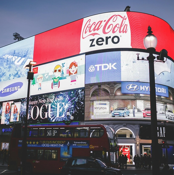 Large digital billboard at night featuring multiple ads including Coca-Cola Zero, Samsung, McDonald's, TDK, Hyundai, and Vogue, above a busy street with a red double-decker bus and people near shop entrances.