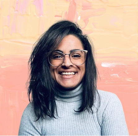 Woman with dark hair and glasses smiling, wearing a light gray ribbed turtleneck sweater.