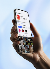 Hand holding a smartphone displaying an Instagram profile page with a red logo, follower and post counts, story highlights, and photo grid visible against a soft blue background.