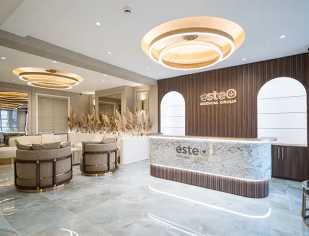 Modern medical clinic reception area with a white marble front desk displaying the Este Medical Group logo, wood-paneled wall behind with matching logo, circular ceiling lights, cushioned round chairs, and neutral-toned decor including pampas grass.
