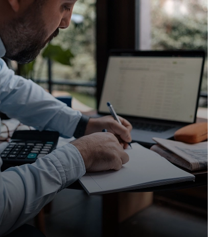 Man in a long-sleeve shirt writing in a notebook at a desk with an open laptop, calculator, and documents nearby.
