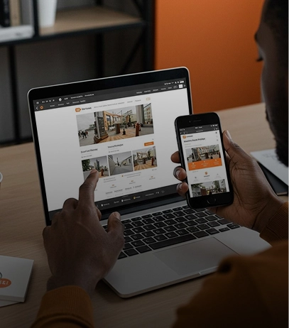 Person using a laptop and holding a smartphone, both displaying a real estate website with property listings and images.