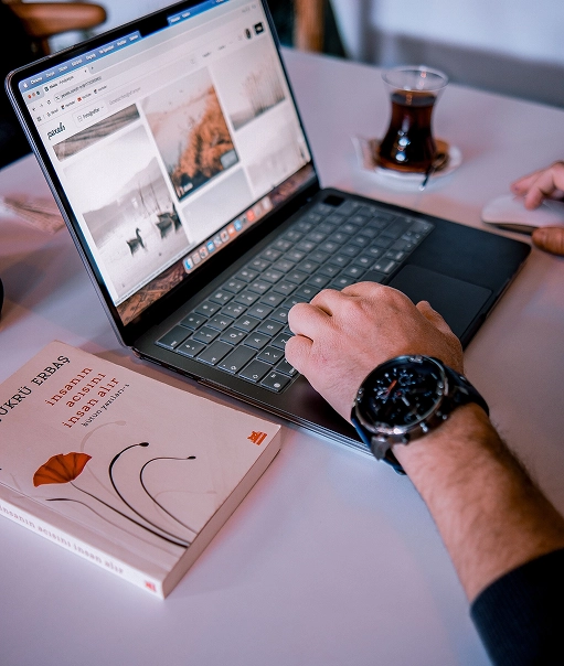 Person using a laptop displaying a webpage with images, seated at a table with a book and a glass of tea nearby. The person's right hand is on the keyboard and the left hand is on a computer mouse, wearing a black watch.