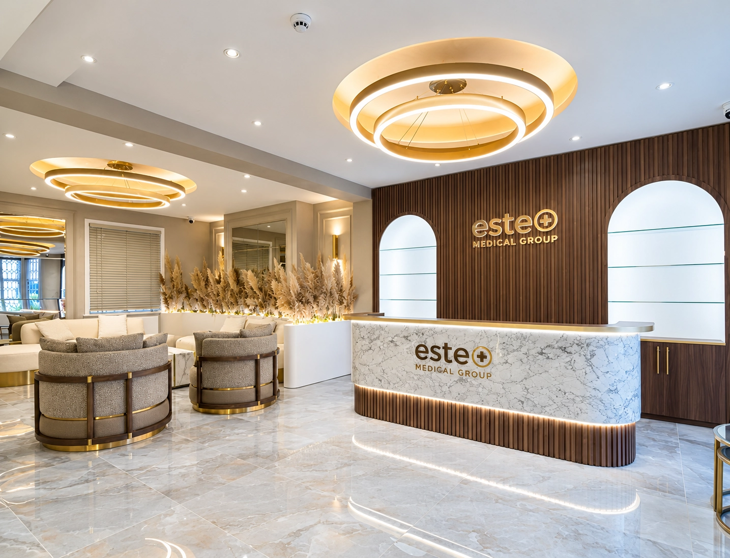 Modern medical office lobby with a marble reception desk branded with 'este+ Medical Group,' wood-paneled wall behind, circular ceiling lights, marble floor, and seating area with beige and gray chairs, white sofa, and decorative plants.