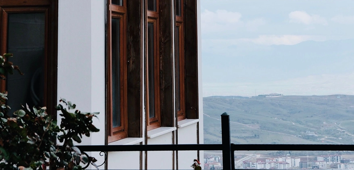 View from a balcony with a black railing showing part of a white building with wooden-framed windows, leafy plants in the foreground, and a distant landscape of hills and buildings under a cloudy sky.