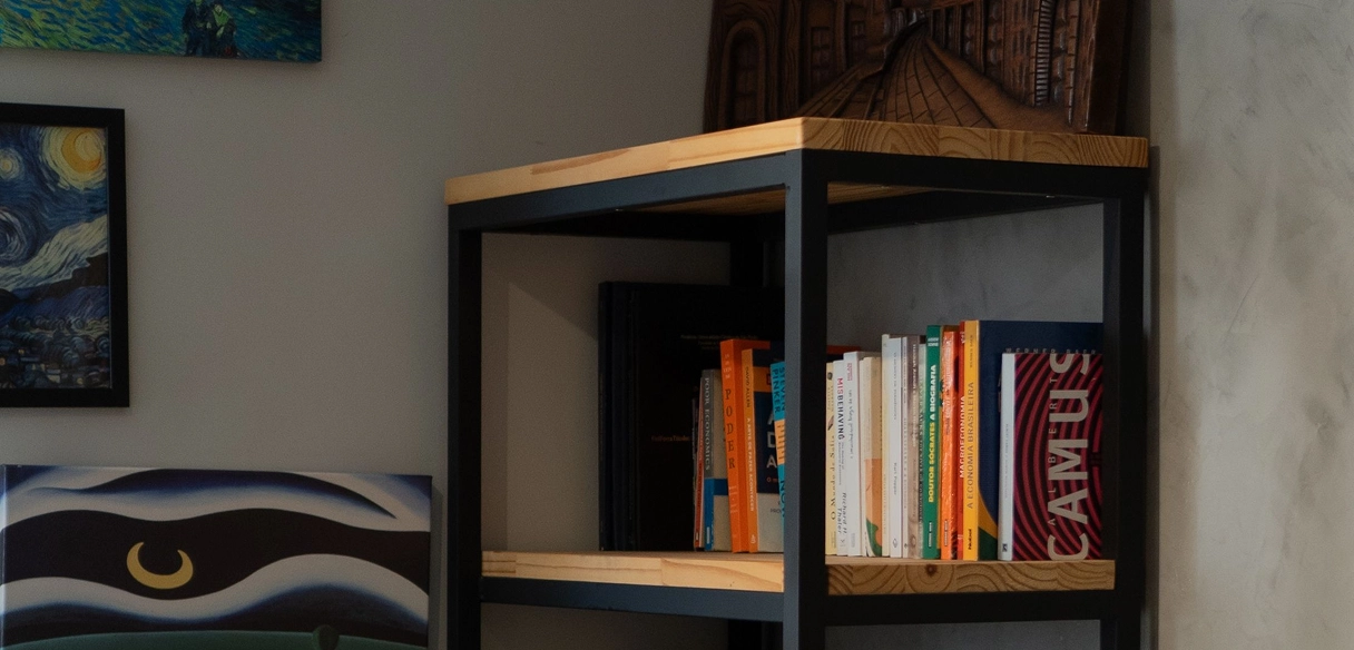 A black metal and wood bookshelf against a light gray wall holds an assortment of books, including a noticeable pink book titled CAMUS on the right side. Part of a painting with a yellow crescent moon is visible on the left side of the image.