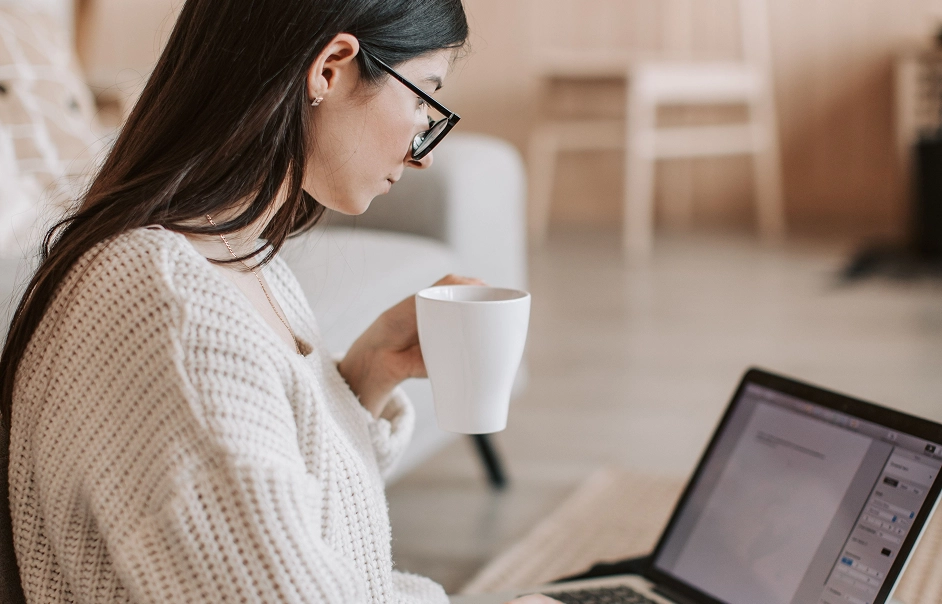 Woman with long dark hair and glasses wearing a white knitted sweater, holding a white mug and looking at a laptop screen indoors.