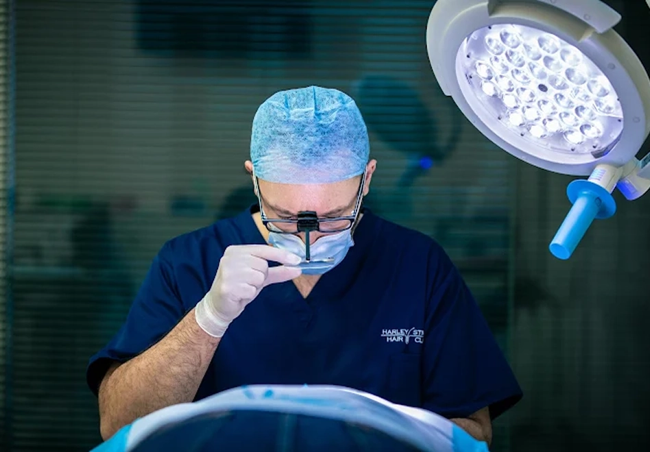 Surgeon wearing blue scrubs, a surgical cap, gloves, and magnifying glasses, focused on performing a procedure under a bright surgical light.