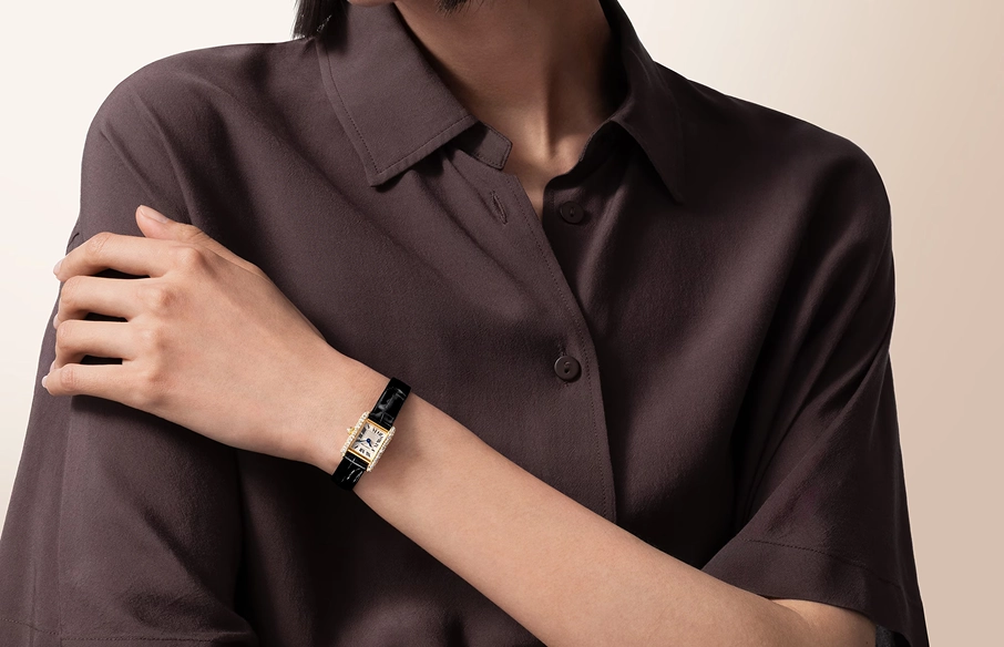 Person wearing a dark brown collared shirt showing a wristwatch with a rectangular gold case and black leather strap.