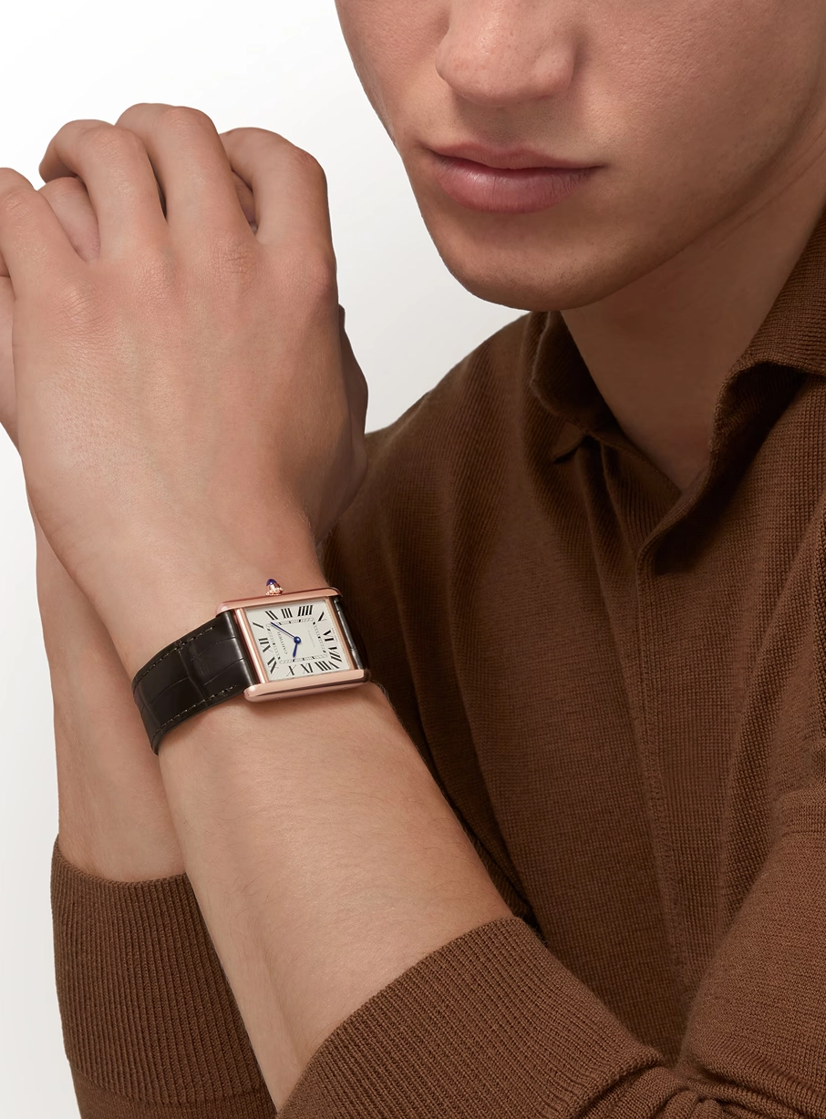 Close-up of a person's wrist wearing a rectangular gold watch with a white dial, black Roman numerals, blue hands, and a black leather strap, dressed in a brown knit sweater.