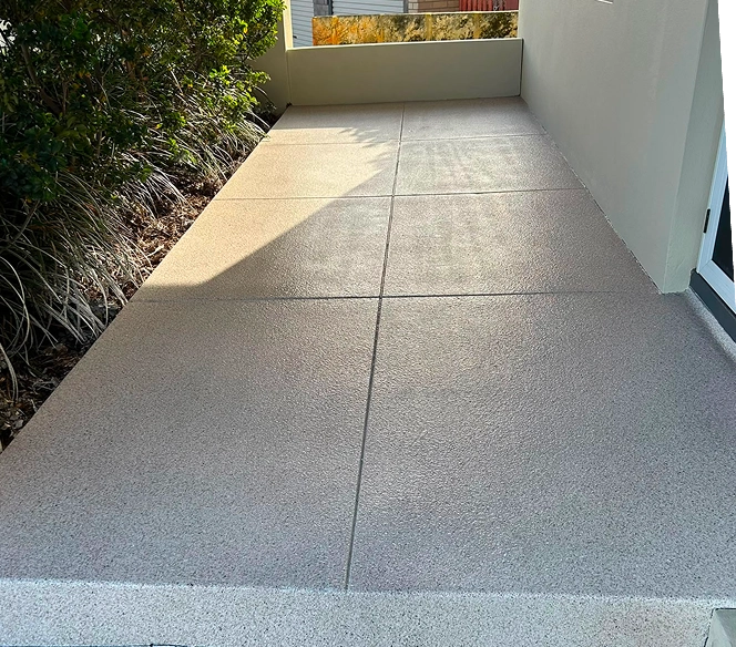 Concrete patio with a textured surface divided into rectangular sections, bordered by green shrubs on the left and a white wall on the right, partially shaded by sunlight.